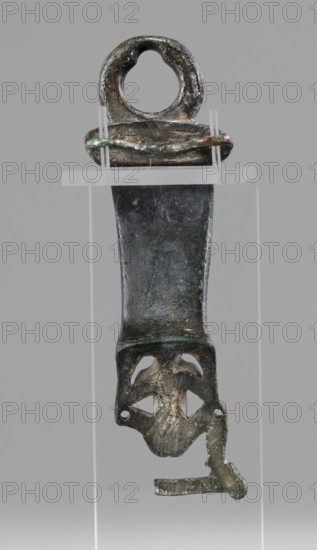 Situla handle with a depiction of Silenus, restitution provenance, Silenus mythology, bronze vessel, 1st half of the 5th century BC, Etruria, vessel, height 13.7 cm, width 4.2 cm, An elongated metallic object with a rounded top featuring a circular hole, mounted on a clear rectangular stand with two vertical supports, showing an overall dark gray and greenish patina with areas of brownish corrosion; the central section is a broad, slightly tapered strip with a textured surface and irregular edges, leading to a lower decorative section with cut-out shapes resembling curved openings and a projecting irregular element, all displaying mottled green, dark gray, brown, and lighter silvery highlights against a pale gray background.