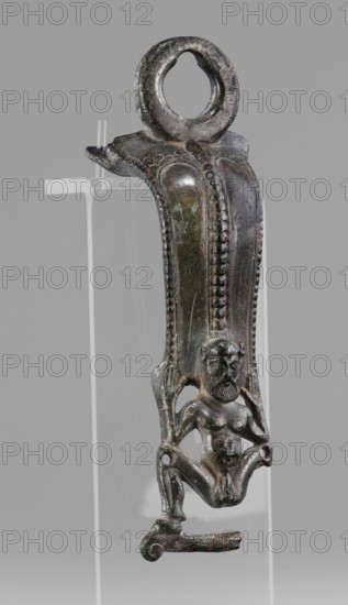 Situla handle with a depiction of Silenus, Silenus mythology, bronze vessel, 1st half of the 5th century BC, Etruria, vessel, height 13.7 cm, width 4.2 cm, The image shows a single metal object with a vertical elongated shape and a circular loop at the top, mounted on a clear stand against a neutral gray background; the metal surface is dark with visible areas of greenish and brownish patina and some lighter metallic highlights, and the object features a relief of a small seated human figure with a bearded face and detailed hair, arms raised beside the head, a defined torso and bent legs with one leg attached to a short curved projection at the bottom, while the sides of the elongated section are decorated with raised bead-like ridges and smooth panels.