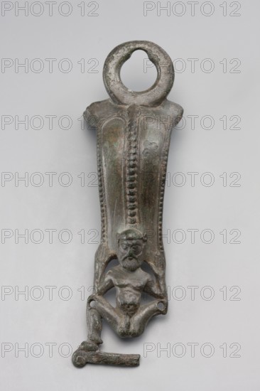 Situla handle with a depiction of Silenus;, Silenus mythology; bronze vessel handle; 1st half of the 5th century BC; Etruria; vessel; height 13.7 cm, width 4.2 cm., A vertical metal object with a rounded ring at the top showing a worn surface and mottled greenish-brown patina, a tapered central shaft with raised beaded ridges and textured panels, and a small seated human figure near the lower end with a beard, defined facial features, muscular torso, and bent legs that connect to a short horizontal bar; overall colors include green, brown, and dark gray.