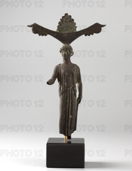 Mirror support with a depiction of Aphrodite. Aphrodite myth., Greek deity. Disc support for a standing mirror; figurine of Aphrodite wearing a Doric chiton. Ca. 450 BC, Sicyon Sykyonia. Hand mirror, mirror figurine. Height 15.5 cm., A small bronze figurine of a standing person mounted on a dark rectangular base, shown against a light gray background; the figure wears a long draped garment with visible folds, has short wavy hair, and one arm extended forward while the other rests at the side; perched atop the figure's head is a horizontal decorative element shaped like a wide V with curled ends and a scalloped piece rising in the center; the object shows areas of greenish patina and overall dark brown metal tones, with the base appearing black and the background light gray.