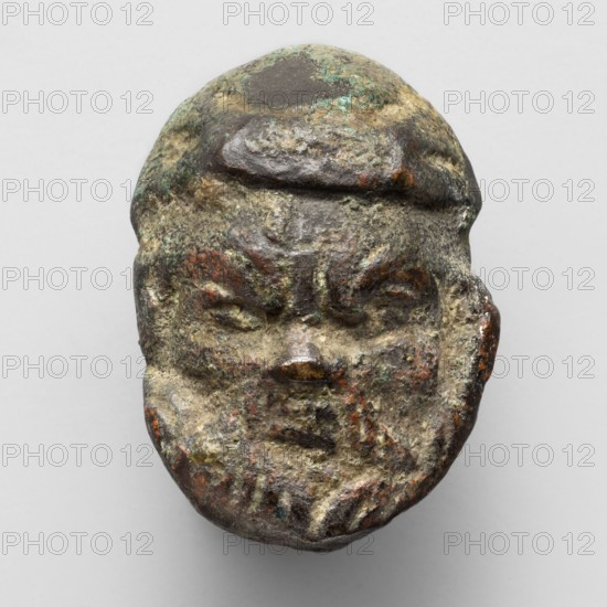 Appliqué in the shape of Silenus's head, The head of a bearded Silenus wearing a cap, designed on one side, with a recess on the opposite side. The round cap, with two diagonal folds on top, running from the forehead to the sides, is pulled slightly over the slightly receding forehead with four vertical wrinkles above the nose. The eyes, set under prominent eyebrows, are deep-set and very narrow, with only the upper, thick eyelids visible. The nose is very short, sunken at the base, wide, and flattened at the nostrils. The small mouth with straight lips, the lower slightly undercut, is framed by two wide strands of mustache, which connect to the closely trimmed facial hair, marked by a thickened surface and several incisions. The slim, sunken cheeks have prominent cheekbones. 4th–3rd century BC, Etruria. Appliqué; height 3.2 cm, width 2.3 cm, depth 1.2 cm; weight 0.046 kg., A worn sculpted head with a rounded top and stylized facial features, showing a brow ridge, closed eyes, a nose, ...