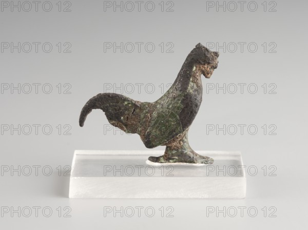 Mirror decoration, 1st half of the 5th century BC, Corinth, mirror, height 3 cm, width 3.6 cm, A small sculptural object of a bird shape stands on a transparent rectangular base; the bird has a curved tail, an upright neck, and a textured surface showing areas of dark brown, green, and hints of lighter brown, with rough, uneven edges and patches of corrosion; the base is translucent and pale, and the background is a smooth, light gray.