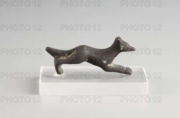 Mirror decoration, 1st half of the 5th century BC, Corinth; mirror; height 1.7 cm, width 3.8 cm., A small dark brown sculptural object shaped like a running animal is shown resting on a clear rectangular base; the object has a rough, pitted surface with several patches of green and lighter brown discoloration, a pointed snout, an arched back, and extended front and rear limbs, all set against a pale gray background.