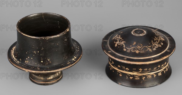 Black-finned pyxis decorated with vine, Greek ceramics, plant motifs, 275–250 BC, Athens, Attica, pyxis vessel, weight 0.16 kg, height 8.3 cm, diameter 8.5 cm, The image shows two round, low pedestal containers with lids placed on a neutral gray surface: both objects are predominantly black with worn areas revealing a warm, light brown or tan underlayer, and one piece features decorative curving vine and grape-like motifs in a lighter brownish-gold tone around its lid and body; the surfaces exhibit scratches, chips, and scattered speckles of the lighter color, and the shapes include flared rims, cylindrical bodies, and circular bases with subtle concentric rings.
