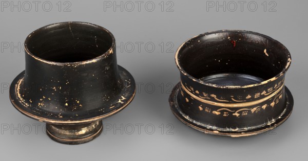 Black-finned pyxis decorated with vine, Greek ceramics, plant motifs, 275–250 BC, Athens, Attica, pyxis vessel, weight 0.16 kg, height 8.3 cm, diameter 8.5 cm., Two shallow cylindrical vessels sit on a smooth gray surface, each with a flared rim and pedestal base; both are primarily black with worn areas revealing a tan or beige underlayer, and one of them displays decorative bands of tan floral and vine motifs around its exterior. The left vessel has a wider flared top with scattered tan speckles and edge wear, a slightly tapered body, and a short rounded pedestal that shows patched tan wear, while the right vessel has a more upright cylindrical profile with two horizontal tan bands and a pattern of small floral shapes and curved lines, along with chipped edges and visible interior wear. The background is a uniform mid-gray.