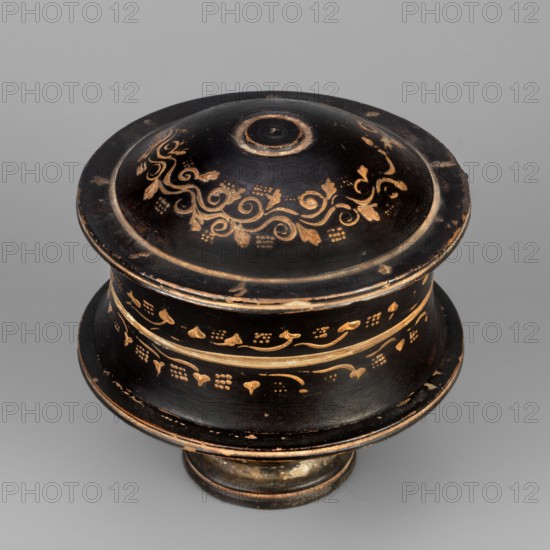 Black-finned pyxis decorated with vine, Greek ceramics, plant motifs, 275–250 BC, Athens, Attica; pyxis vessel; weight 0.16 kg, height 8.3 cm, diameter 8.5 cm., A round lidded container with a domed top and a flared body, shown from above on a neutral gray background. The object is predominantly black with decorative worn gold-colored patterns and bands around the lid and body, including scroll-like motifs and small floral or dot cluster accents. The rim and base show exposed lighter areas where the black surface is chipped or rubbed, revealing a brownish or tan underlayer. The lid has a raised central knob with concentric rings, and the overall surface shows a matte to slightly worn sheen. Colors visible include black, gold, brown, tan, and gray.