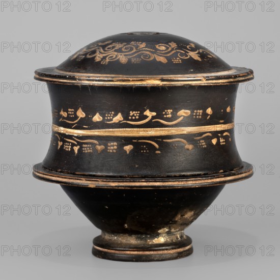 Black-finned pyxis decorated with vine, Greek ceramics, plant motifs, 275–250 BC, Athens, Attica; pyxis vessel, weight 0.16 kg, height 8.3 cm, diameter 8.5 cm., A rounded lidded vessel with a bulbous body and flared rim, shown against a neutral gray background. The object is predominantly black with decorative metallic gold painted motifs forming bands and scrolling patterns around the lid and upper and middle sections. The lid is domed and sits on a slightly overhanging rim, with visible wear where the black surface has been scuffed to reveal lighter underlayers. The body narrows toward a small foot, which shows areas of discoloration and surface abrasion in brownish and off-white tones. Overall the visible colors are black, metallic gold, brown, off-white, and gray.