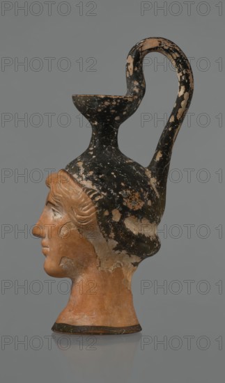 Oinochoe in the shape of a woman's head, Greece, Attica, ca. 450 BC. Oinochoe vessel fashioned from yellowish clay with an orange surface and, in places, shiny black engobe; some elements painted white. The upper part and the headdress are covered with black engobe, on which a white olive wreath stands out. The neck is outlined at the base with a black band. The elongated female face, very carefully modeled, corresponds to the classic Attic head type from the second half of the 5th century. The oval eyes are surrounded by a black outline. The hair is divided by a parting and falls in waves to ear level. The headdress flows into a narrow neck ending in a wide, cup-shaped spout. A cylindrical handle is attached vertically between the rim and the back of the head. Preservation condition: missing engobe in some places, damaged surface. Iconography: head; culture: Greek., The image shows a sculpted pottery vessel in the shape of a human head viewed in profile against a plain gray background...