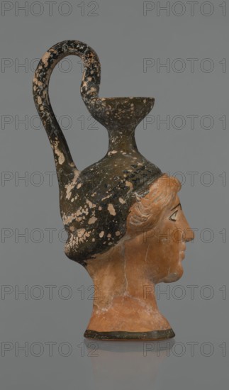 Oinochoe in the shape of a woman's head. Yellowish clay with orange surface and areas of shiny black engobe; some elements painted white. The upper part and headdress are covered with black engobe, with a white olive wreath on the headdress. The neck is outlined at the base with a black band. The elongated female face, very carefully modeled, corresponds to the classic Attic head type from the second half of the 5th century. Oval eyes are surrounded by a black outline. Hair is parted and falls in waves to ear level. The headdress flows into a narrow neck ending in a wide, cup-shaped spout. A cylindrical handle vice is attached vertically between the rim and the back of the head. Preservation: missing engobe in places, damaged surface. Ca. 450 BC, Attica. Oinochoe vessel., The image shows a sculpted head-shaped vessel photographed in profile against a plain gray background, with the face turned to the right. The object combines a terracotta-colored human head with a dark vessel forming ...
