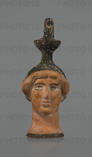 Oinochoe in the shape of a woman's head. Yellowish clay with orange surface, in places shiny black engobe; some elements painted white. The upper part and the headdress are covered with black engobe, with a white olive wreath on the headdress. The neck is outlined at the base with a black band. The elongated female face, very carefully modeled, corresponds to the classic Attic head type from the second half of the 5th century BC. Oval eyes are surrounded by a black outline. Hair is divided by a parting and falls in waves to ear level. The headdress flows into a narrow neck ending in a wide, cup-shaped spout. A cylindrical handle is attached vertically between the rim and the back of the head. Preservation: missing engobe in some places, damaged surface. ca. 450 BC, Attica. Oinochoe vessel, head iconography, Greek., The image shows a terracotta sculpted head of a person with stylized wavy hair and a long neck, topped by a tall, spotted black object that resembles a handle or vessel feat...