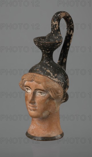 Oinochoe in the shape of a woman's head, Greece, ca. 450 BC, Attica. Oinochoe vessel in the shape of a woman's head, yellowish clay with orange surface and in places shiny black engobe; some elements painted white. The upper part and the headdress are covered with black engobe, the headdress bearing a white olive wreath. The neck is outlined at the base with a black band. The elongated female face, very carefully modeled, corresponds to the classic Attic head type from the second half of the 5th century. The oval eyes are surrounded by a black outline. The hair is divided by a parting and falls in waves to ear level. The headdress flows into a narrow neck ending in a wide, cup-shaped spout. A cylindrical handle/vice is attached vertically between the rim and the back of the head. Preservation condition: missing engobe in some places; damaged surface. Iconography: head; culture: Greek; ancient vessel; oinochoe., A sculpted terracotta female head with stylized wavy hair and a calm facial...
