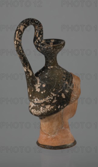 Oinochoe in the shape of a woman's head, repossession Bytom. Greece. Oinochoe, head iconography, Greek culture, ancient vessel. Yellowish clay, orange surface, in places shiny black engobe. Some elements painted white. The upper part is covered with black engobe, as is the headdress, on which a white olive wreath stands out. The neck is outlined at the base with a black band. The elongated female face, very carefully modeled, corresponds to the classic Attic head type from the second half of the 5th century. The oval eyes are surrounded by a black outline. The hair is divided into two parts by a parting and falls in waves to ear level. The headdress flows into a narrow neck ending in a wide, cup-shaped spout. A cylindrical vice is attached vertically between the rim and the back of the head. Preservation condition: missing engobe in some places, damaged surface. Ca. 450 BC, Attica, oinochoe vessel., A photographed ancient pottery vessel shaped like a head with a tall, curved handle and...
