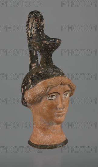 Oinochoe in the shape of a woman's head, Greece, Attica, ca. 450 BC. Oinochoe vessel fashioned from yellowish clay with an orange surface, in places covered with shiny black engobe; some elements painted white. The upper part and the headdress are covered with black engobe, on the headdress a white olive wreath is depicted. The neck is outlined at the base with a black band. The elongated female face, very carefully modeled, corresponds to the classic Attic head type from the second half of the 5th century. The oval eyes are surrounded by a black outline. Hair is parted and falls in waves to ear level. The headdress flows into a narrow neck ending in a wide, cup-shaped spout. A cylindrical handle vice is attached vertically between the rim and the back of the head. Preservation condition: missing engobe in some places; damaged surface. Iconography: head. Culture: Greek., A photographed terracotta head-shaped vessel with a long neck and a curved handle forming a spout, showing a human f...