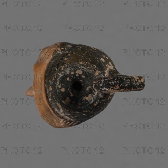 Oinochoe in the shape of a woman's head, Greece, ca. 450 BC, Attica. Oinochoe wine jug shaped as an elongated female head, classic Attic head type from the second half of the 5th century. Yellowish clay with orange surface and, in places, shiny black engobe; some elements painted white. Upper part and headdress covered with black engobe; a white olive wreath on the headdress. Neck outlined at the base with a black band. Oval eyes outlined in black; hair parted and falling in waves to ear level. Headdress flows into a narrow neck ending in a wide, cup-shaped spout. A cylindrical vice handle attachment is attached vertically between the rim and the back of the head. Preservation condition: missing engobe in some places, damaged surface., A worn ceramic vessel photographed from above showing a round opening and a curved handle, with a mottled surface in black and tan and areas of exposed terracotta brown; a rim and decorative flaring at one side in a natural clay brown; the background is ...