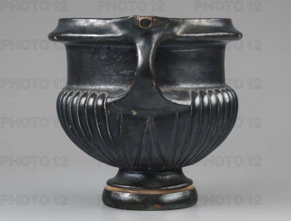 Kantharos, unknown Attic workshop, 320–310 BC, Attica. Kantharos vessel, weight 0.692 kg. The vessel has a high echinus-shaped foot, modelled with a single recess at three-quarters of its height. Rounded, fluted body under the vises the recesses take an oblique direction, creating a three-leaf pattern; on one side the side vises are ajar due to lack of space, narrow shoulders, and a high smooth neck. The vises are strongly rounded. Modelled spout with a strip below the rim, and characteristic horizontal, elongated, triangular, slightly raised handles above the vises. The bottom features an ornament of six 13-leaf palmettes, not entirely symmetrically arranged; above them is a roulette-like decoration six rows. The entire vessel, including the inner surface of the foot, is covered with varnish except for the edge of the foot on which it stands and a single depression on the foot. Technology: Varnish is shiny, rather metallic, distributed evenly over the surface. Preservation: a few chip...