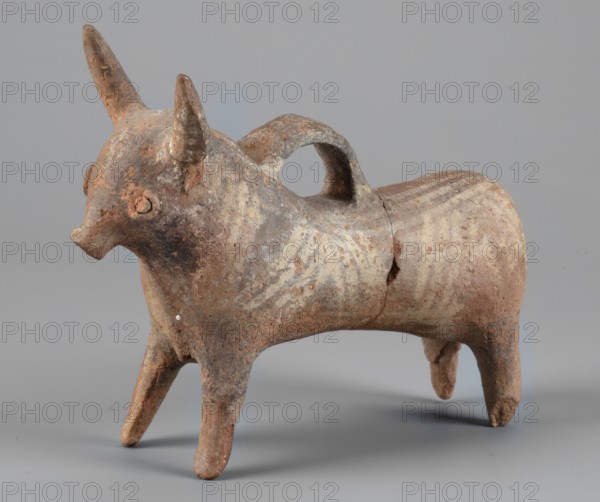 Bull-shaped vessel. Vessel in the shape of a bull standing on widely spaced legs. The cylindrical body, straight cut, flows into a wide neck. The head is small with a cylindrical muzzle, with plastically crafted eyes and horns protruding upwards. There is a hole in the neck, and beneath it is a vice, the other end of which is attached in the middle of the back. The surface is covered with geometrically painted decoration. Cypriot pottery, askos. 1300–1200 BC, Cyprus. Height 11 cm; width 13 cm; depth 4.5 cm; weight 0.11 kg., A small sculptural object shaped like an animal with a rounded body, four legs, a short tail, and two upright horn-like projections on its head sits against a neutral background. The surface shows a weathered, rough texture with areas of worn patina and flaking. The overall color palette includes brown, tan, beige, and gray, with patches of darker brown and some lighter cream tones forming faint, irregular patterns on the body. A curved handle-like form rises from t...