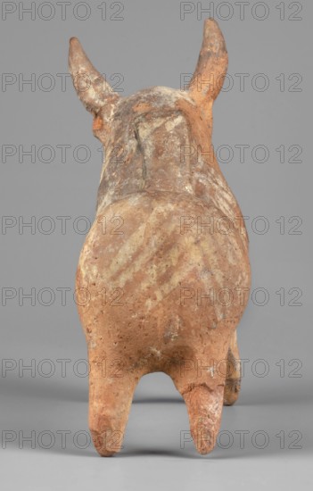 Bull-shaped vessel, Vessel in the shape of a bull standing on widely spaced legs. The cylindrical body, straight cut, flows into a wide neck. The head is small with a cylindrical muzzle, with plastically crafted eyes and horns protruding upwards. There is a hole in the neck, and beneath it is a vice, the other end of which is attached in the middle of the back. The surface is covered with geometrically painted decoration. 1300–1200 BC, Cyprus. Ceramic vessel askos. Height 11 cm, width 13 cm, depth 4.5 cm, weight 0.11 kg. Bulls; animal figurines; Cypriot pottery., A rear view of a sculpted animal figure with a rounded body, two upward-pointing horns, and four legs, showing a weathered surface with rough texture and areas of flaking; the piece displays a mix of earthy browns, reddish-browns, pale tan patches, and small hints of gray and white against a neutral gray background.