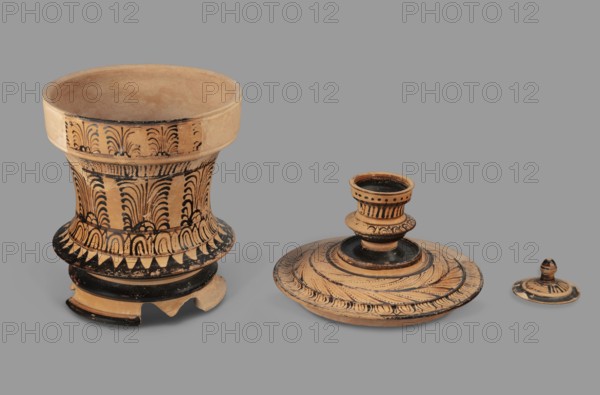 Pyxis with a lid, unknown Boeotian workshop. Pyxis with a lid with a high handle. Cylindrical body with slightly concave walls. Decorated with a plant motif. Set on three legs. Sharp edge above the leg. Bottom decorated with a black circle, with a varnish stain in the central part. Technology: visible traces of turning; good-quality glossy varnish; cleaned clay. Notes: reconstructed artefact, large part of the rim and body of the vessel and lid restored with plaster additions. Maker/attribution:, date: 2nd half of the 4th century BC; place of origin: Boeotia; type: pyxis vessel; weight: 0.950 kg., A group of three ceramic objects sits on a flat surface against a gray background: a tall, flared vessel on the left with a wide rim and a decorated body showing repeating fan-like and arch patterns in black on a tan clay, a low, circular object in the center with a raised central cup-like structure and concentric decorative bands and brushstroke patterns in black on tan, and a small lid-like...