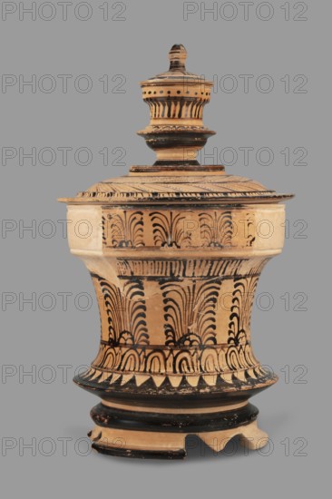 Pyxis with a lid, unknown Boeotian workshop. Pyksis with a lid with a high handle. Cylindrical body with slightly concave walls. Decorated with a plant motif. Set on three legs. Sharp edge above the leg. Bottom decorated with a black circle, with a varnish stain in the central part. Technology: visible traces of turning, good quality glossy varnish, cleaned clay. Notes: reconstructed artefact, large part of the rim and body of the vessel and lid, plaster additions. Maker attribution/author unknown. 2nd half of the 4th century BC, Boeotia. Type: pyxis vessel. Weight 0.950 kg., A ceramic lidded vessel with a rounded base and a tall urn-like top, shown against a plain gray background, featuring a beige or light tan body decorated with black painted ornamental patterns including repeated curved and feather-like motifs, bands, and triangular shapes; the lid has concentric horizontal ridges and dotted and striped black accents, and the base shows chipped areas and wear with some missing frag...