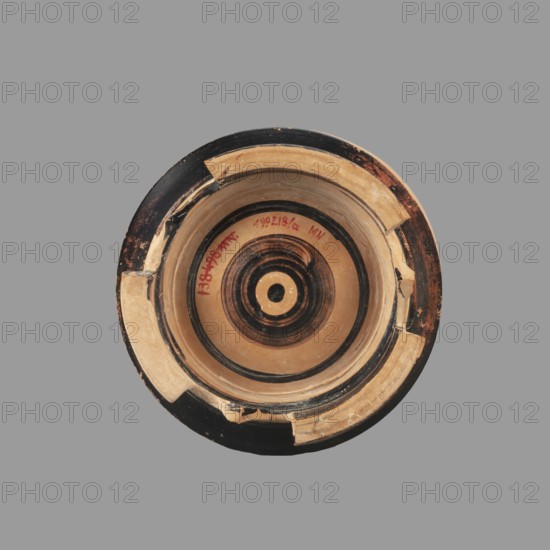 Pyxis with a lid, unknown Boeotian workshop. Pyxis with a lid with a high handle. Cylindrical body with slightly concave walls. Decorated with a plant motif. Set on three legs; there is a sharp edge above the leg. The bottom is decorated with a black circle with a varnish stain in the central part. Technology: visible traces of turning; good quality glossy varnish; cleaned clay. Notes: reconstructed artifact—large part of the rim and body of the vessel and lid have plaster additions. 2nd half of the 4th century BC, Boeotia. Pyxis, vessel; weight 0.950 kg., A circular ceramic object viewed from above with concentric rings and a central circular motif, showing areas of tan, black, and dark brown; the rim has chipped and repaired sections with lighter tan patches and visible wear, and the background is a uniform gray.