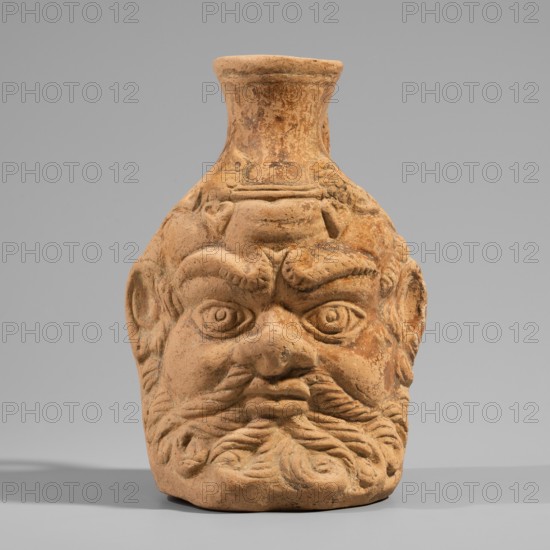 Vessel with a belly in the shape of a satyr's head, unknown Pergamon workshop. A flat-bottomed vessel with a plastic belly in the shape of a Silenus's head, a funnel-shaped spout, and a vertical ribbon handle curved into a circle. The Silenus's head has horse ears and an ivy wreath with flowers above the forehead. The face has a low, wavy cola, small eyes with thin, slender eyelids and marked irises and pupils, a short, broad nose, chubby cheeks, and a narrow, closed mouth. A significant part of the face is covered by a beard and mustache, parted in the middle and arranged in twisted locks with curled ends. Orange clay, 2nd century, Pergamon; vessel. Height 13.6 cm, width 9.6 cm, depth 10.5 cm, weight 0.246 kg., A terracotta vessel shaped like a stylized human head with a pronounced brow, deep-set round eyes, a broad nose, and a full curled beard; the surface shows textured sculpted details and worn areas. The object sits on a flat surface and casts a soft shadow. The background is a s...