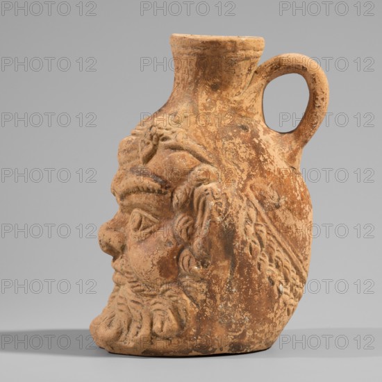 Vessel with a belly in the shape of a satyr's Silenus's head. A flat-bottomed vessel with a plastic belly in the shape of a Silenus's head, a funnel-shaped spout, and a vertical ribbon handle curved into a circle. The Silenus's head has horse ears and an ivy wreath with flowers above the forehead. The face has low, wavy hair, small eyes with thin, slender eyelids and marked irises and pupils, a short broad nose, chubby cheeks, and a narrow closed mouth. A large part of the face is covered by a beard and mustache, parted in the middle and arranged in twisted locks with curled ends. Orange clay, Roman pottery, 2nd century, Pergamon workshop, Vessel. Height 13.6 cm; width 9.6 cm; depth 10.5 cm; weight 0.246 kg., A weathered terracotta jug shaped like a stylized human head with a rounded handle near the top, showing sculpted facial features including a prominent nose, brows, eyes, ear, and a detailed beard and hair, with textured worn surfaces and patches of roughness; the visible colors a...
