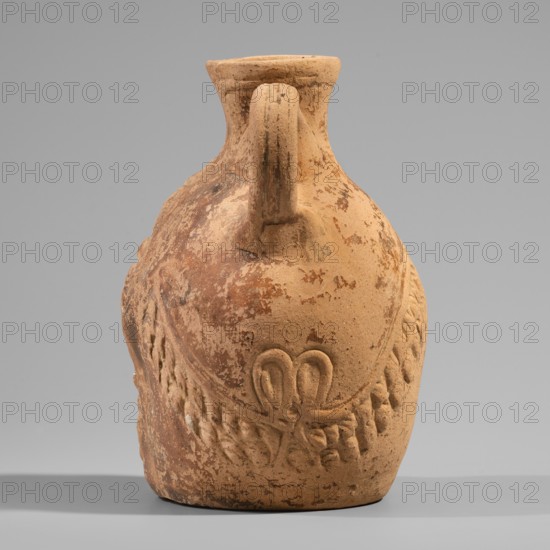 Vessel with a belly in the shape of a satyr's Silenus's head, unknown Pergamon workshop. A flat-bottomed Roman pottery vessel with a plastic belly in the shape of a Silenus's head, a funnel-shaped spout, and a vertical ribbon handle curved into a circle. The Silenus's head has horse ears and an ivy wreath with flowers above the forehead. The face has a low, wavy coiffure, small eyes with thin, slender eyelids and marked irises and pupils, a short broad nose, chubby cheeks, and a narrow closed mouth. Much of the face is covered by a beard and mustache parted in the middle and arranged in twisted locks with curled ends. Orange clay, 2nd century, Pergamon; vessel; H 13.6 cm, W 9.6 cm, D 10.5 cm; weight 0.246 kg., A weathered clay jug sits against a plain gray background, showing a rounded body with a narrow neck and a single sturdy handle that arches from the rim to the shoulder. The surface shows irregular patches of worn and flaking material, revealing variations of warm tan, light brow...
