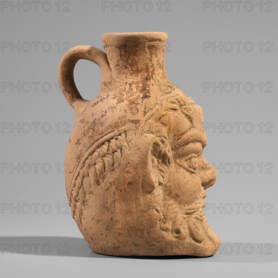 Vessel with a belly in the shape of a satyr's Silenus's head. A flat-bottomed Roman pottery vessel with a plastic belly in the shape of a Silenus's head, a funnel-shaped spout, and a vertical ribbon handle curved into a circle. The Silenus's head has horse ears and an ivy wreath with flowers above the forehead. The face has a low, wavy chiton — note: original said cola likely chignon? — small eyes with thin, slender eyelids and marked irises and pupils, a short, broad nose, chubby cheeks, and a narrow, closed mouth. Much of the face is covered by a beard and mustache, parted in the middle and arranged in twisted locks with curled ends. Orange clay, 2nd century, from Pergamon. Vessel; height 13.6 cm, width 9.6 cm, depth 10.5 cm, weight 0.246 kg., A terracotta jug shaped like a human head with a handle emerging from the back, showing a sculpted face in profile with pronounced facial features including an eye, nose, mustache, beard, and textured hair; the surface is weathered with rough p...