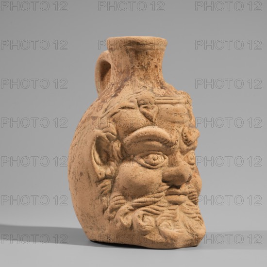 Vessel with a belly in the shape of a satyr's head, unknown Pergamon workshop. A flat-bottomed vessel with a plastic belly in the shape of a Silenus's head, a funnel-shaped spout, and a vertical ribbon handle, curved into a circle. A Silenus's head with horse ears and an ivy wreath with flowers above the forehead. The face has a low, wavy chignon, small eyes with thin, slender eyelids and marked irises and pupils, a short, broad nose, chubby cheeks, and a narrow, closed mouth. A significant part of the face is covered by a beard and mustache, parted in the middle and arranged in twisted locks with curled ends. Orange clay, 2nd century, Pergamon, Roman pottery; vessel height 13.6 cm, width 9.6 cm, depth 10.5 cm, weight 0.246 kg., A sculpted terracotta vessel shaped like a bearded human head with a handle at the back, showing a textured surface in shades of light brown and tan with darker brown weathering, pronounced eyebrows and deep-set eyes, a broad nose, stylized mustache and curly b...