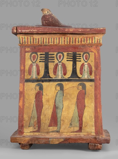 Canopic box with the falcon Sokaris on the roof;, falcons, djed pillars, tit iconography; naos; grave goods; 664–525 BC, Late Period, 26th Dynasty, Egypt; canopic box; height 42 cm, width 22.5 cm, depth 29.5 cm, weight 2.414 kg, A rectangular painted wooden box with a lid topped by a small sculpted bird in brown tones; the box shows a decorative band near the top with vertical striped motifs in yellow, red, and blue, and broad red borders at the sides. The main painted panel is divided horizontally into two registers: the upper register features a series of stylized figures with rounded heads and red and pale green garments flanking tall black vertical shapes, set on a yellow background; the lower register shows a row of standing human figures in profile wearing long red and pale green robes with black hair against a yellow ground. The base and edges of the box show exposed wood in brown and reddish tones, and the overall palette includes yellow, red, black, brown, pale green, and blue...