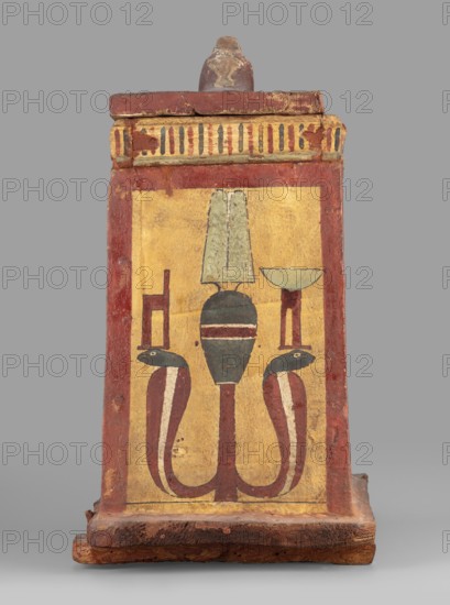 Canopic box with the falcon Sokaris on the roof; unknown creator; falcons, djed pillars, tit iconography; naos; grave goods; 664–525 BC, Late Period, 26th Dynasty, Egypt; canopic box; height 42 cm, width 22.5 cm, depth 29.5 cm, weight 2.414 kg, A painted rectangular wooden box with a slightly raised top and base, shown against a gray background; the front face displays a central stylized motif of a tall vertical element topped by a pale green fan shape, supported by a dark green rounded form with horizontal bands, and flanked by two curved serpent-like forms with dark green heads and red bodies that curve downward and then up; a simple bowl-like shape on a short red stand and a narrow chair-like shape flank the central motif; the painted field is a warm yellow framed by a broad red border, with a decorative band of vertical stripes in red, green, and dark tones along the upper edge; the wood shows worn and chipped areas revealing darker brown beneath, and the overall colors visible inc...