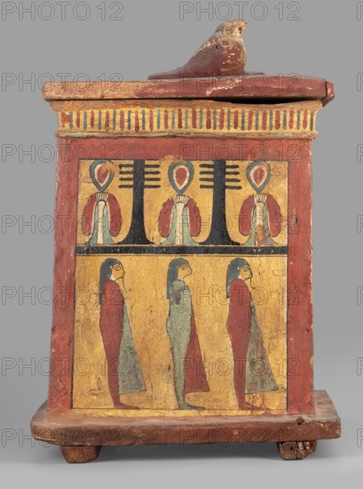 Canopic box with the falcon Sokaris on the roof; falcons; djed pillars iconographic; tit iconographic; naos; grave goods; 664–525 BC, Late Period, 26th Dynasty, Egypt; canopic box; height 42 cm, width 22.5 cm, depth 29.5 cm, weight 2.414 kg;, A rectangular wooden box with a lid topped by a carved bird figure sits on a short base with small feet; the box is painted in muted red, yellow, black, blue-green, and pale green tones. The front face is divided into two horizontal registers bordered by a red frame: the upper register shows a repeated decorative motif with stylized plant-like shapes in yellow and black and teardrop elements in red and blue-green, while the lower register depicts a group of standing human-like figures rendered in profile wearing long garments in red and blue-green against a yellow background. The lid and upper edge display a striped decorative band in alternating yellow and darker hues, and the wooden surfaces show areas of wear and chipped paint.