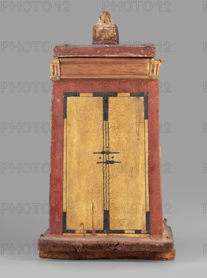 Canopic box with the falcon Sokaris on the roof; falcons, djed pillars, tit iconography; naos; grave goods; 664–525 BC, Late Period, 26th Dynasty, Egypt; canopic box; height 42 cm, width 22.5 cm, depth 29.5 cm, weight 2.414 kg;, A rectangular wooden box with a flat base and a small decorative finial on top, painted in red, yellow, black, and natural wood tones; the front face shows a large yellow panel framed by a red border with black linear accents and a simple black geometric motif near the center; the top section has a natural wood band beneath a red cap and small carved corner blocks with traces of yellow and black; the base is darker, rough-textured wood with visible wear and cracks, and the overall surface shows aged paint, chips, and areas of exposed wood.
