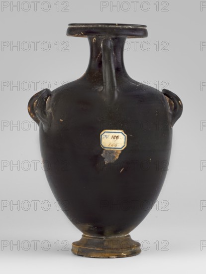 Red-figure hydria with a combat scene, Laghetto-Caivano Group c. 340–320 BC; Duel Painter 340–330 BC. Hydria decorated with a combat scene of two naked hoplite warriors fighting with long spears; they wear pointed helmets, hold round convex shields in their left hands, and wear greaves on their legs. Weaponry painted white with yellow retouching. Ca. 340–320 BC, Campania. Vessel hydria, height 22.5 cm, width 15.6 cm, depth 13.3 cm, weight 0.612 kg., A dark-colored ceramic vase with a narrow neck, flared rim, and rounded body is centered against a light gray background; the vase has a glossy black surface with visible wear showing small areas of chipped or rubbed spots in lighter tan and brown tones, a few looped handles near the shoulder, and a short footed base that reveals worn brownish-gold coloring.