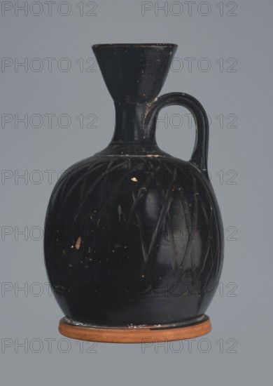 Ariballus lekythos, 4th century BC, Nola findspot, Campania origin, lekythos vessel, height 12.0 cm, width 7.0 cm, A dark vase with a bulbous body, a narrow neck flaring into a wide mouth, and a single curved handle at the shoulder, sitting on a flat round base; the vase surface is predominantly black with visible scuffs and small areas where the dark coating is worn away to reveal a lighter material, and the base ring is a terracotta color; the background is a flat muted gray.