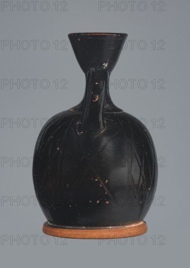 Ariballus lekythos, 4th century BC, Nola, Campania; lekythos vessel, height 12.0 cm, width 7.0 cm., A single ceramic vessel centered against a solid pale blue-gray background, with a rounded black body that shows worn spots revealing a terracotta color beneath, a narrow neck rising to a flared rim, and a single black handle connecting neck and shoulder; the base is exposed terracotta and the surface of the black glaze has a few scratches and chips.