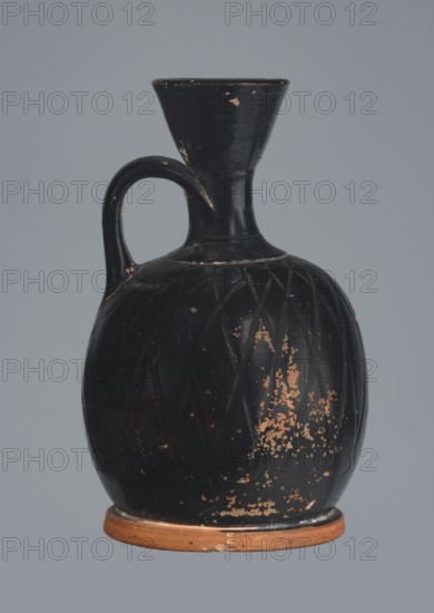 Ariballus lekythos, 4th century BC, Nola, Campania; lekythos vessel, height 12.0 cm, width 7.0 cm., A dark pottery vessel with a rounded body, narrow neck, flared rim, and a single curved handle, shown against a plain light gray background; the vessel is primarily black with areas of worn orange-brown clay visible where the black surface has chipped, and faint incised diagonal patterns on the body; the base rim is predominantly orange-brown and shows small chips and wear.