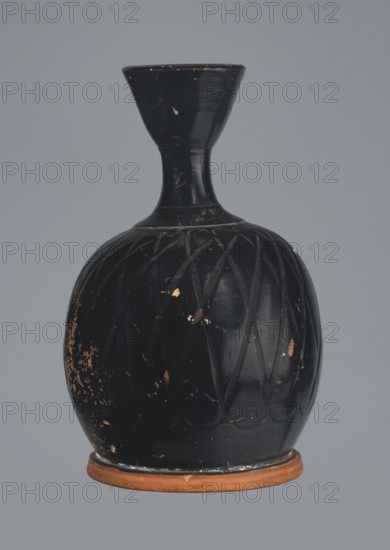 Ariballus lekythos, 4th century BC, Nola, Campania; lekythos vessel, height 12.0 cm, width 7.0 cm., A rounded vase with a narrow neck and flared rim sits against a plain gray background; the vase is predominantly black with a glossy finish and shows a few worn spots revealing a brownish-orange underlayer, a group of shallow incised diagonal patterns around the bulbous body, and a thin exposed brownish-orange base.