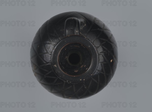 Ariballus lekythos, 4th century BC, Nola, Campania, lekythos vessel, Ariballus lekythos, height 12.0 cm, width 7.0 cm, A top-down view of a round, dark ceramic vessel with a central opening and a small handle, showing a glossy black surface with a carved geometric pattern and areas of worn, lighter brown where the finish has chipped, set against a muted gray background.