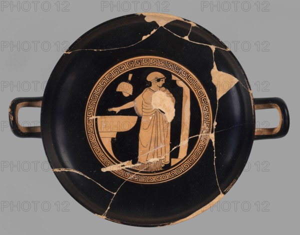 Red-figure kylix with a bathing scene, the Boots Painter ca. 460 BC. Bath iconography: women wash at lutrions bowls on high bases, pass around folded garments, and one ties characteristic high boots. Greek ceramics; Attica/Athens place of origin. Goblet with slightly parted rim edges, shallow bowl, low stem, and flat foot. Vices have a round cross-section, slightly raised tops reaching the line of the rim. Conical bottom. Sleeve-shaped foot. Technology: thick, very good quality varnish, evenly distributed; smooth surface; traces of turning invisible, brush marks visible; mixture: mica. Notes: glued from many fragments; visible conservation restorations; surface contaminated; visible secondary losses and chips; standing surface heavily worn. Kylix; vessel. Dimensions: height 9.2 cm, width 29.8 cm, depth 22.8 cm, weight 0.53 kg., A round ceramic dish with a glossy black surface and a group of pale orange painted figures and decorative bands at its center; the dish has two horizontal hand...