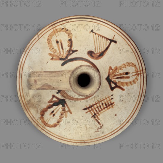 Lagynos with a depiction of musical instruments – lyres and syrinxes, unknown Eastern Greek workshop. Lagynos on a ring-shaped foot, a belly separated by a sharp edge from the convex arms, and a cylindrical neck terminating in a ring-shaped rim. The vise connecting the arms to the upper part of the neck is profiled. The belly, covered with white paint, is decorated with orange lines running all around. Similar lines decorate the arms near the belly. On the shoulders there are depictions of three wreaths and musical instruments lyres and syrinxes painted with brown paint. 2nd century BC, Greece; found in Cyprus. Lagynos vessel, weight 0.41 kg, height 16 cm, diameter 16.5 cm., A circular ceramic object seen from above with a central round opening and an attached handle, predominantly cream colored with brown decorative brushwork; the surface shows a few concentric brown rings near the edge and several brown painted motifs including curved lines, a grid-like mark, and leaf-shaped or wreat...