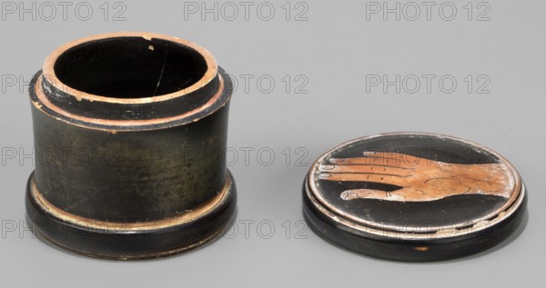 Red-figure pyxis with an image of a hand on the lid. Small pyxis with a lid. Cylindrical shape, separated foot, concave bottom. Flat lid with slightly protruding edges. Rounded sides. Depicts a hand on the lid. Toilet vessel, Greek ceramic. 5th century BC, Attica possibly Arsinoe/Athens., Technology: Glossy, high-quality varnish, evenly distributed but unevenly fired; green shades visible in places. Smooth surface; cleaned clay. Traces of turning visible under the varnish. Admixtures: mica. Condition notes: Intact. White deposit visible on inside surface of the lid. Secondary losses and abrasions visible. Weight: 0.10 kg., A cylindrical container with its lid placed beside it occupies a neutral background; the container and lid are primarily black with visible worn areas showing a brownish-orange underlayer, and the interior of the cylinder appears dark. The lid features a depiction of a hand in brownish-orange against the black surface with some lighter worn spots, and both pieces sho...