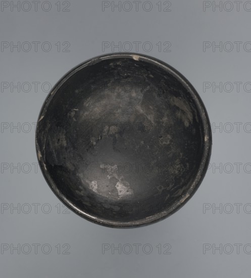 Bucchero Nero cup, Goluchów Greater Poland Voivodeship, Etruria Italy, 6th century BC, vessel, height 5.1 cm, width 6.0 cm, A round, shallow bowl viewed from above with an interior surface that is predominantly dark gray to black, showing areas of lighter gray wear and patches of a muted beige where the finish is worn; the rim is slightly uneven and shows small chips of lighter color; the background is a smooth pale gray.