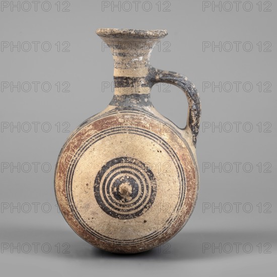 Barrel-shaped jug, unknown Cypriot workshop, geometric motifs, Cypriot ceramics. A vessel with a barrel-shaped body terminated on both sides by small protrusions. The cylindrical neck terminates in a cup-shaped spout. A band-shaped vise connects the body to the neck midway up. The decoration, finished with brown paint, consists of concentric bands encircling the barrel. Two wide light brown bands on either side of the vise are framed on either side by narrow dark brown bands. At the protrusions, the bands group into a set of concentric circles. Transverse bands between two vertical bands decorate the vise. A series of concentric belts on the neck. 850–600 BC, Cyprus, Arsinoe. Vessel; height 12 cm, width 10 cm., A round, flat-bodied ceramic vessel with a narrow neck and flared rim, featuring a curved handle attached from the neck to the shoulder; the surface shows concentric circular painted bands on the body and circular target-like rings at center, with areas of paint wear and surface...