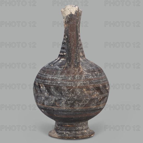 Oinochoe, unknown Cypriot workshop; Arsinoe Cyprus. Oinochoe on a cup-shaped foot with a spherical body transitioning into sloping arms, separated by a recess from the narrow, tapering neck terminating in a triangular rim. The band-shaped handle protrudes slightly above the rim and is connected to the arm. The vessel is covered with gray-blue, matte paint. Decoration in white and brown paint surrounds the vessel concentrically. On the foot, stripes of white paint decorate the body; a laurel sprig adorns the narrowest part of the body, with white and brown stripes above it. On the shoulders, a ray ornament extends up to the neck. On the neck at the base and under the rim, white dots, and between the rows of dots, white stripes. 332–30 BC; Hellenistic period, Cyprus; oinochoe vessel; height 14 cm, width 9 cm., An aged rounded vessel with a narrow tall neck and a flared foot is shown against a plain gray background; the surface displays a weathered, mottled pattern in dark brown and black...