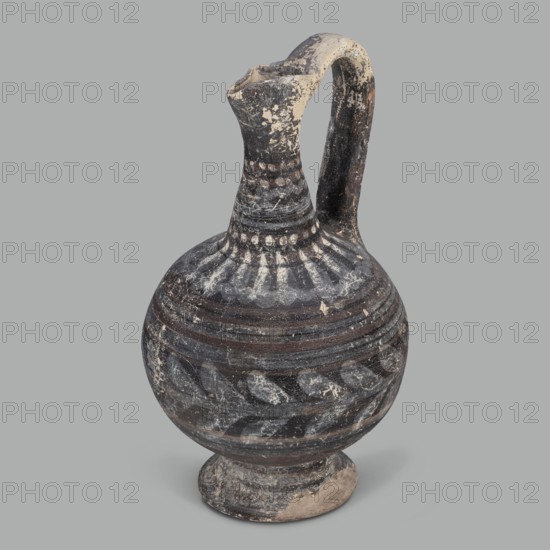 Oinochoe, unknown Cypriot workshop; Arsinoe Cyprus. Oinochoe on a cup-shaped foot with a spherical body transitioning into sloping arms, separated by a recess from the narrow, tapering neck terminating in a triangular rim. The band-shaped handle protrudes slightly above the rim and is connected to the arm. The vessel is covered with gray-blue, matte paint. Decoration in white and brown paint surrounds the vessel concentrically. On the foot, stripes of white paint decorate the body; a laurel sprig adorns the narrowest part of the body, with white and brown stripes above it. On the shoulders, a ray ornament extends up to the neck. On the neck at the base and under the rim, white dots, and between the rows of dots, white stripes. 332–30 BC; Hellenistic period, Cyprus. Oinochoe vessel. Height 14 cm, width 9 cm., A single rounded ceramic jug with a narrow neck, flared lip, and a single curved handle rising from the neck to the shoulder, resting on a small circular foot; the surface shows wo...
