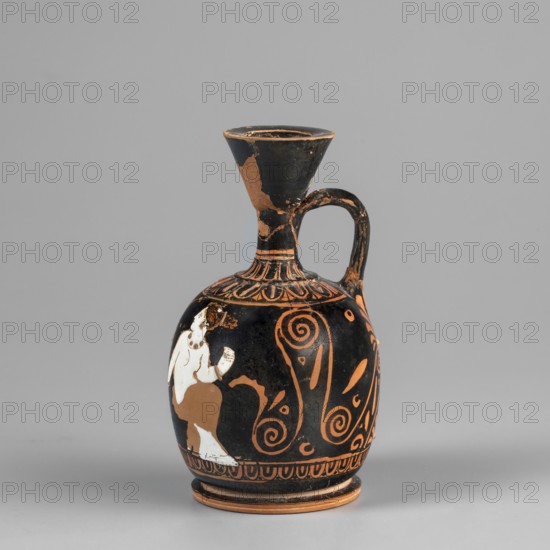 An aryballus lekythos on a ring-shaped, profiled foot, with a bulging, egg-shaped belly, convex handles flowing into a neck terminating in a cup-shaped rim. The handle zone is narrow and band-like. At the base of the neck is a bar ornament, and on the handles is an egg-shaped motif with plastic lines in ovals. Below the handle zone are two upward-facing palmettes, arranged one above the other, decorated with leaves and volutes. The front of the vessel depicts a seated, nude woman wearing a garment covering her legs and hips; she faces left with her head turned right. Behind her is a leaning, winged Eros, standing on his right leg with his left leg raised, holding a necklace in his outstretched hands. The composition is framed from below by an egg-shaped motif. Preservation condition: the neck, handle, and rim are glued; a few chips are visible on the surface. ca. 420 BC earlier dating: 4th century BC, Apulia. Vessel type: lekythos/aryballus lekythos. Height 9.4 cm; width 5.0 cm; foot d...