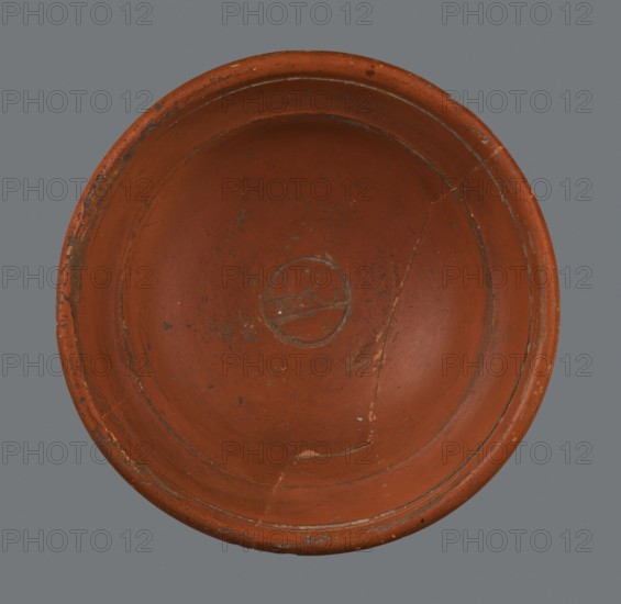 Terra sigillata bowl, Nasso Workshop; Dragendorff 1895 form 27. Fine-grained clay, orange-brown 2.5YR 5/8; brown-red glaze 2.5YR, gives a delicate shine and covers the entire surface of the vessel. Whole vessel, glued from two fragments; late 1st–early 2nd century AD 130–180 AD, Lavoye; height 6.5 cm, diameter 13.9 cm rim., A round shallow dish viewed from above with a worn reddish-brown surface and a slightly raised rim, showing a few areas of darker wear and small spots of lighter abrasion; there are visible cracks and repaired joins along the interior, a faint circular impression near the center, and the background is a muted gray.