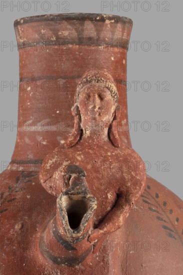 Jug with a figurine of a woman with a jug. Cypriot pottery, female figurines applique. Jug on a foot resembling an inverted echinus with a belly widening by convex shoulders, separated from a cylindrical neck with a straight spout. A two-part vise connects the neck with the shoulders, and on the side opposite the vise is the bust of a woman with a jug. Decoration executed in white and black paint: the belly at its widest point and along one-third of its height is decorated with alternating white and black concentric bands. Beneath the vise, a wavy stripe of black paint extends onto the shoulders on both sides. On both sides, the shoulders feature two birds among plants, a row of dots and a concentric stripe at the base of the neck; a branch with leaves encircles the neck above, and black triangles on a white background at the edge. Dated 480–400 BC; Cypriot — Classical Period I. Arsinoe Marion provenance. Vessel; height 35 cm, width 19 cm, weight 1.72 kg., A close-up view of a reddish-...