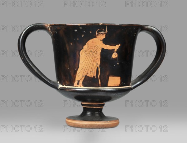 Red-figure kantharos with a depiction of an ephebe with an aryballos and a Greek inscription “Beautiful boy”., Kantharos with a slightly flared rim, a deep bowl, a flat bottom, a high profiled stem, ring-shaped foot; thin-walled. A: bearded man with a lyre in his head in left profile, holding a lyre in his left hand. B: ephebe with aryballos. Inner surface of the bowl coated with varnish. Technique: smooth surface; varnish thick, shiny, unevenly fired. Condition: vessel intact; small secondary losses and chips on surface. Varnish color: 10 YR 2/1 black. Circa 450 BC mid-5th century BC. Attica provenance. Type: kantharos vessel. Dimensions: height 11 cm; width 16.2 cm; depth 10.2 cm., A two-handled ceramic vessel with a rounded bowl and a short stem rests against a neutral background; the vessel has a glossy black surface with orange-red figures and details, showing a standing robed figure in profile reaching toward a small round object held in the other hand, and a rectangular block ne...
